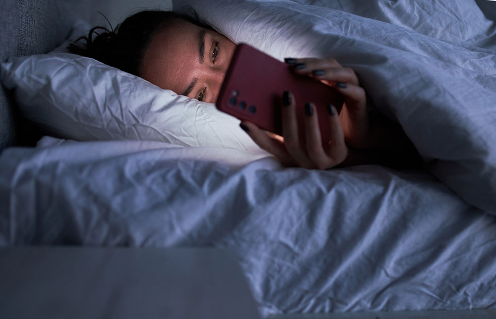 Young woman lies on her side in bed at night, holding a phone up close to her face with both hands