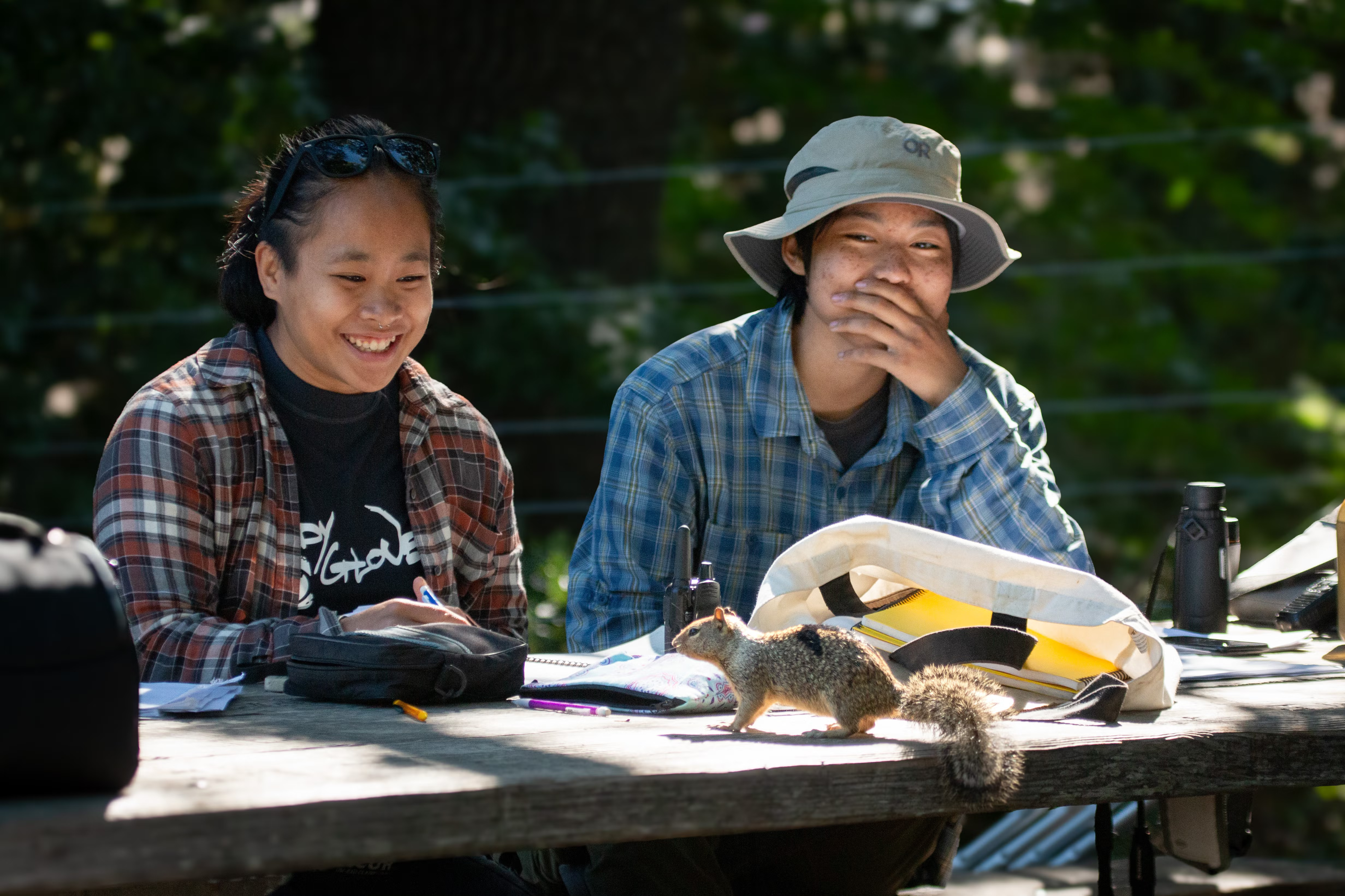 Two young undergraduate researchers smile at a squirrel that's jumped on a picnic table, one with her hand over her mouth