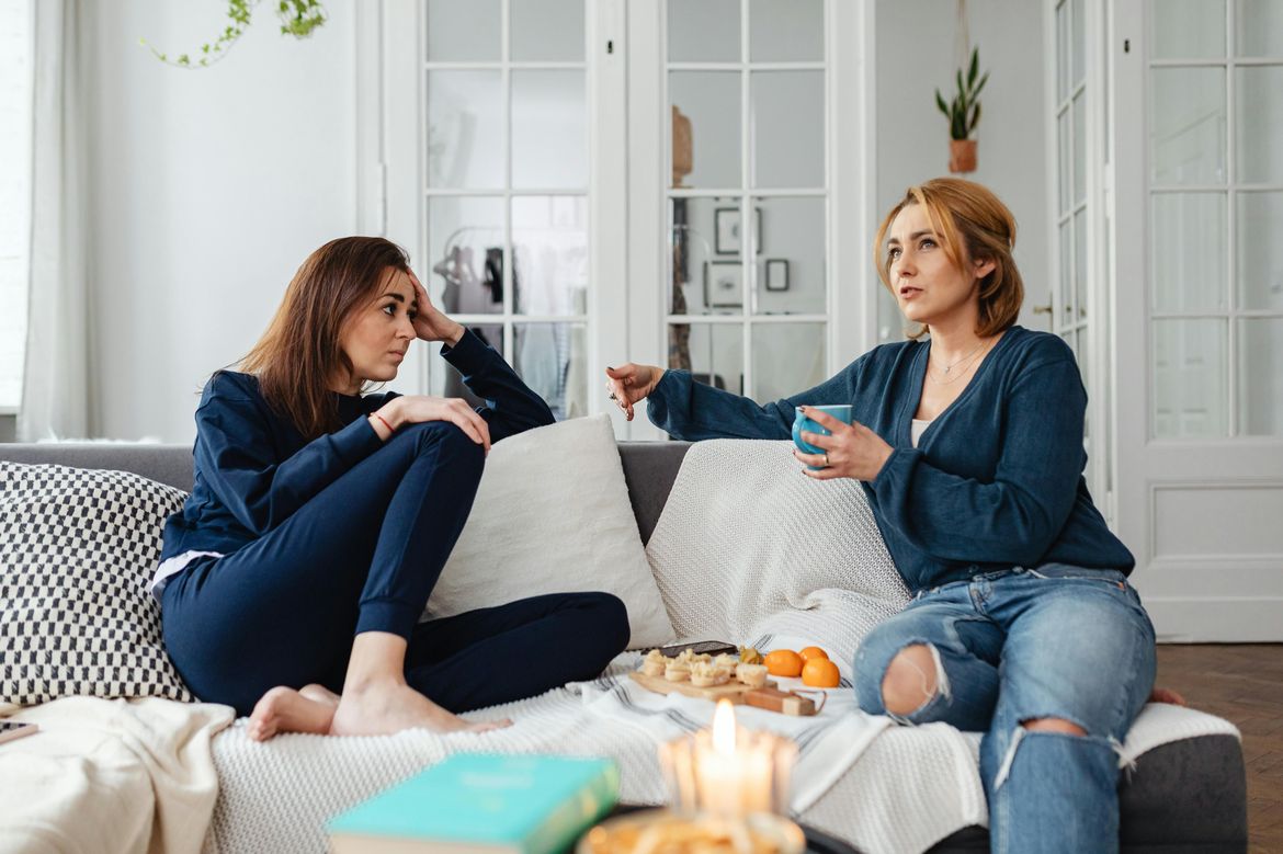 Two women drink tea and talk to each other on a couch in an airy white living room