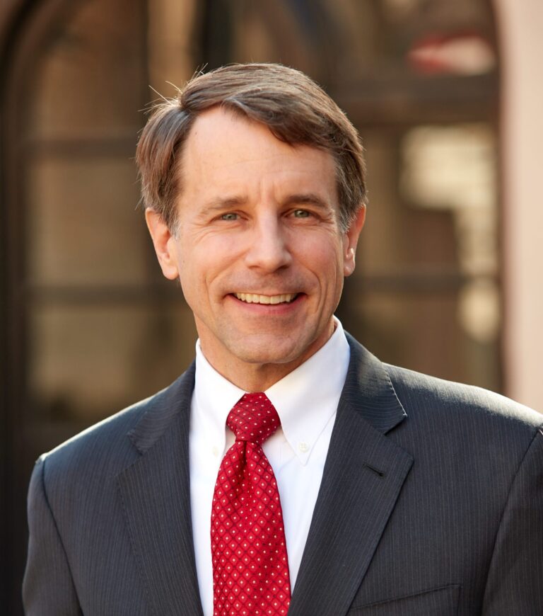 Dave Jones smiles for a headshot wearing a dark suit and red tie