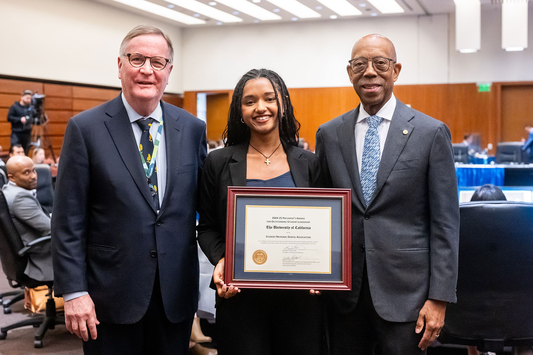 Betty Birbo holds a plaque and smiles for a photo with UCSF Chancellor Sam Hawgood and UC President Michael V. Drake.