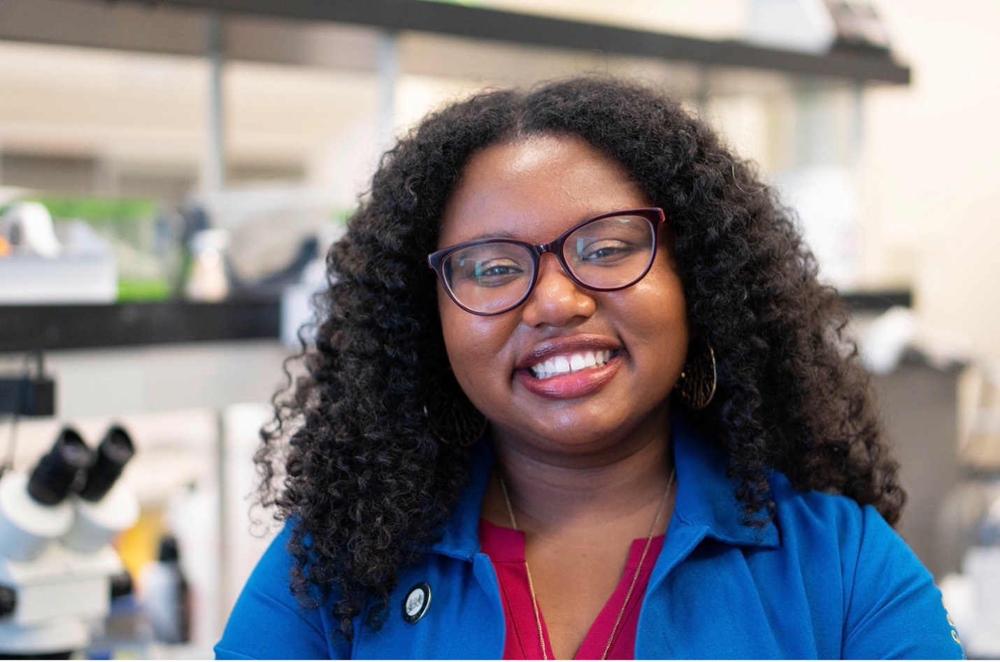 Young woman with curly hair and glasses in blue lab coat smiling