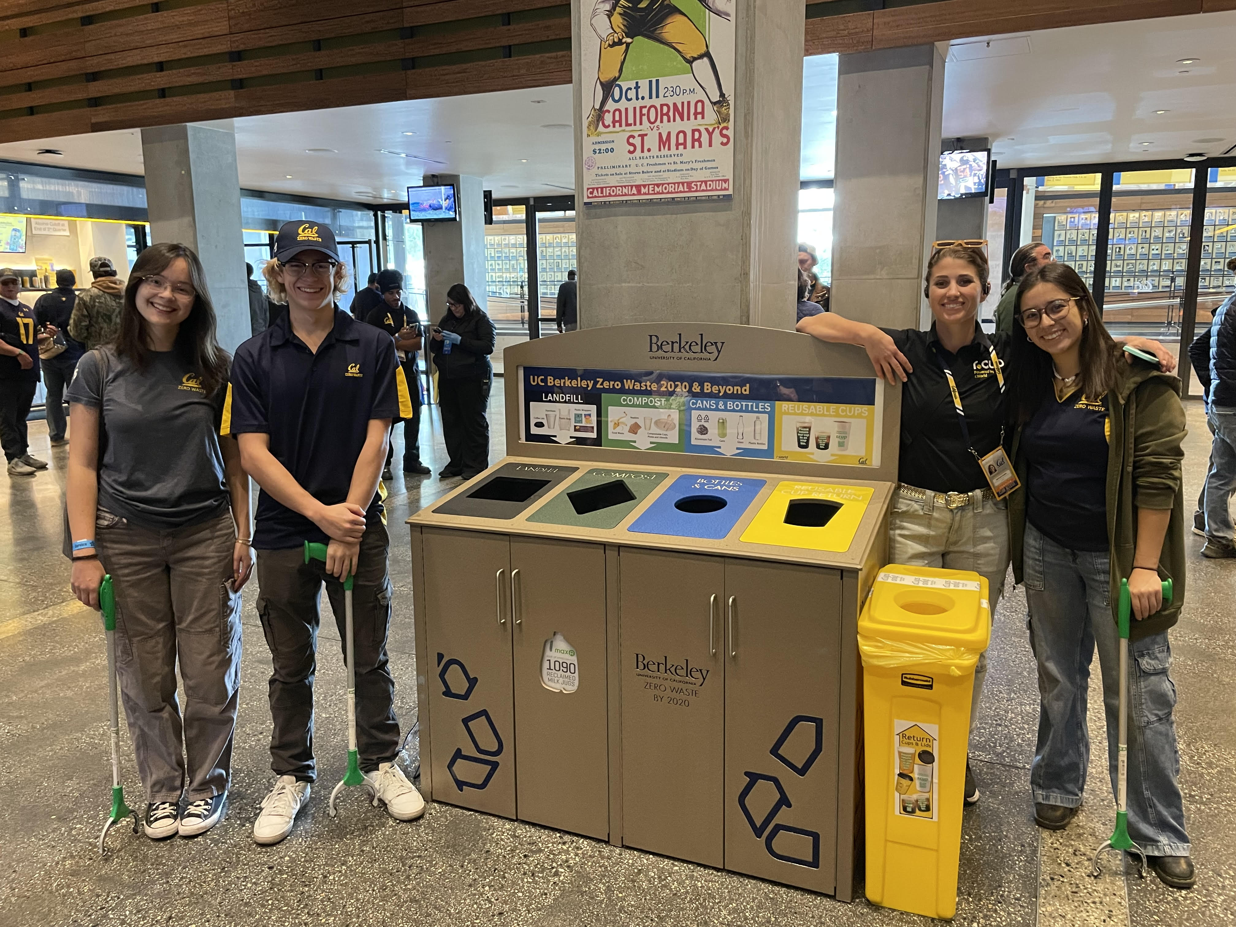 Four students wearing Cal polo shirts and holding trash pickers stand next to waste disposal bins at Cal Memorial Stadium