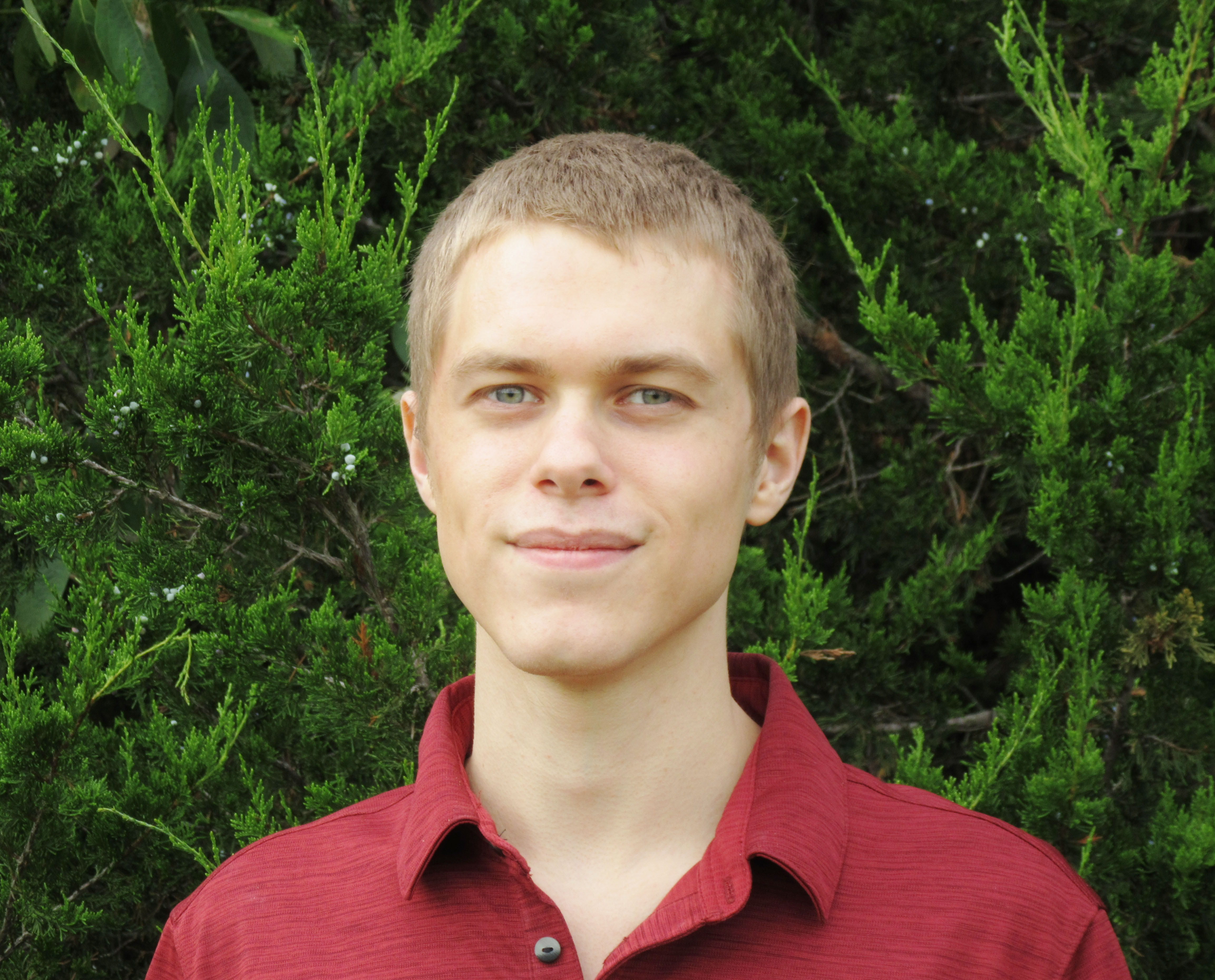 Young man with short hair and a red shirt photographed in front of greenery