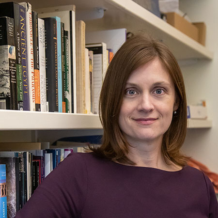 Woman with shoulder-length brown bob and a purple shirt looks at camera in front of bookshelves