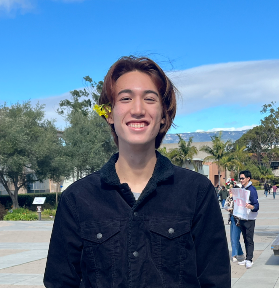 Jake Twomey smiles at the camera wearing a yellow flower tucked behind his ear.