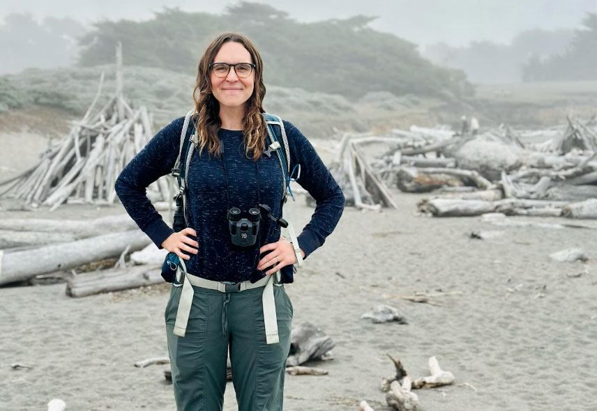 Michelle Fullner smiles for the camera with hands on her hips, standing on a foggy beach with lots of driftwood behind her.