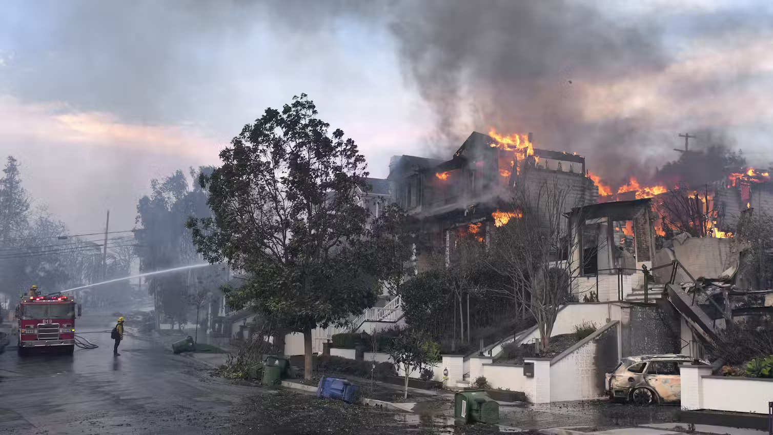 The upper levels of a home on fire in a nice neighborhood, a fire truck spraying the home with water from a hose while a firefighter looks on