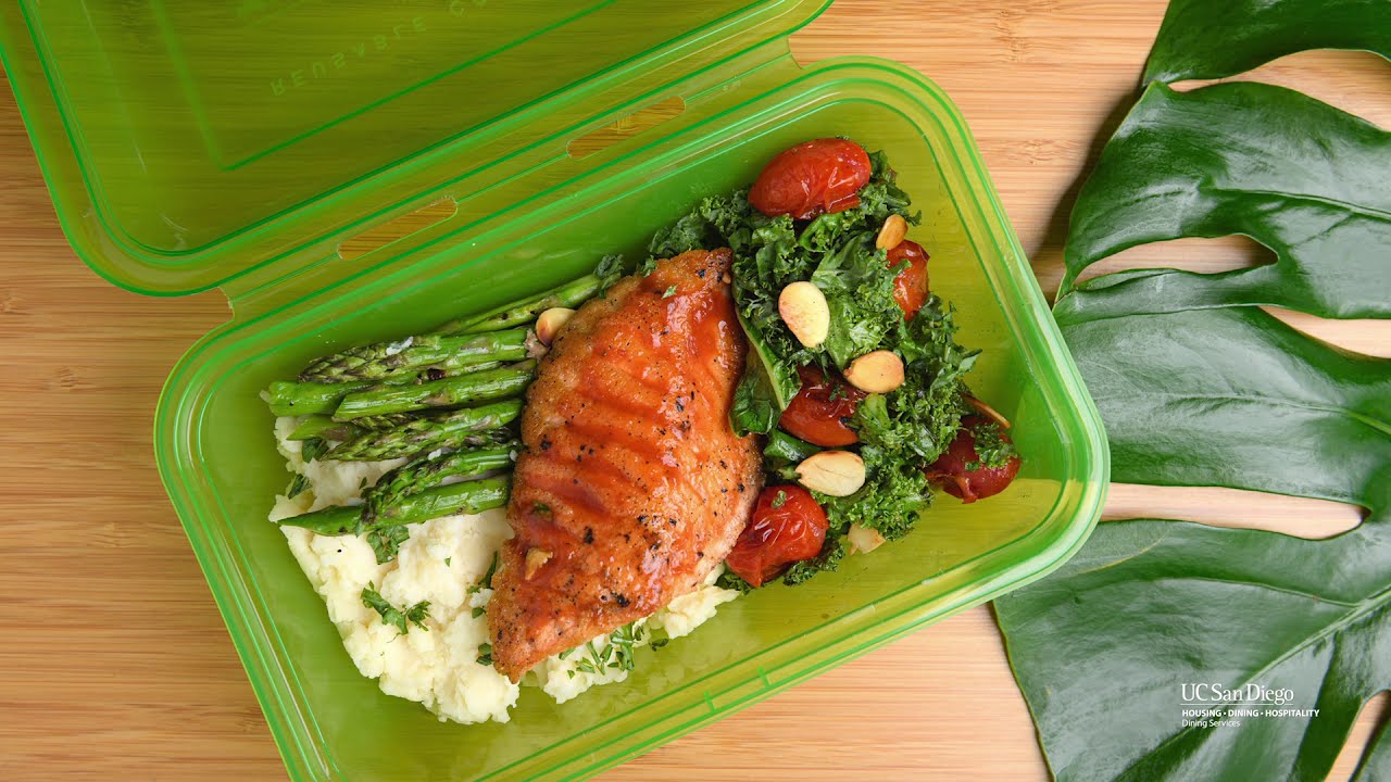 A green plastic takeout container open to show a meal of rice, asparagus, salad and chicken.