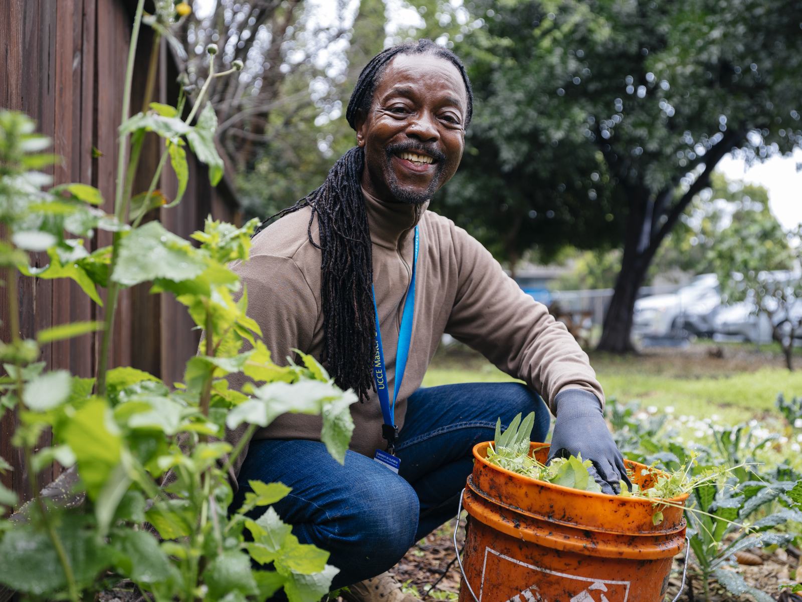 A man with long braids pauses from weeding a garden to smile for a photo