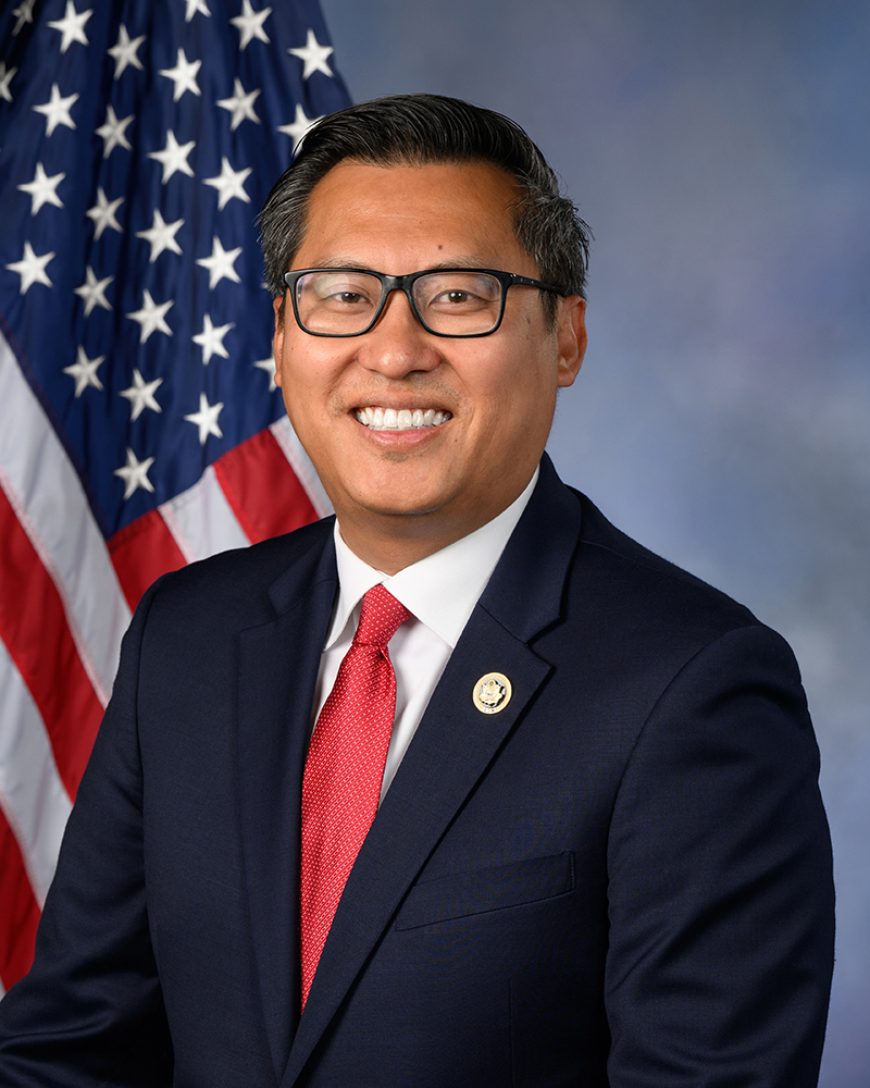 Representative Vince Fong, wearing a suit and a red tie, smiles for a portrait in front of an American flag.