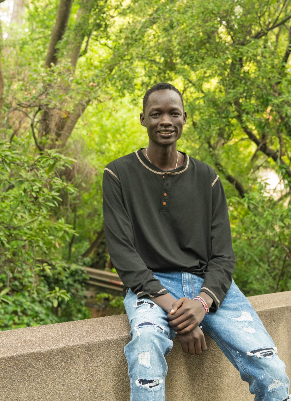 Man in sweater and jeans smiles at camera, sitting on a supporting wall with trees behind him on the Berkeley campus