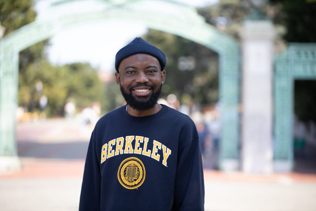 Young man in a blue cap and a Berkeley sweatshirt poses in front of Sather Gate