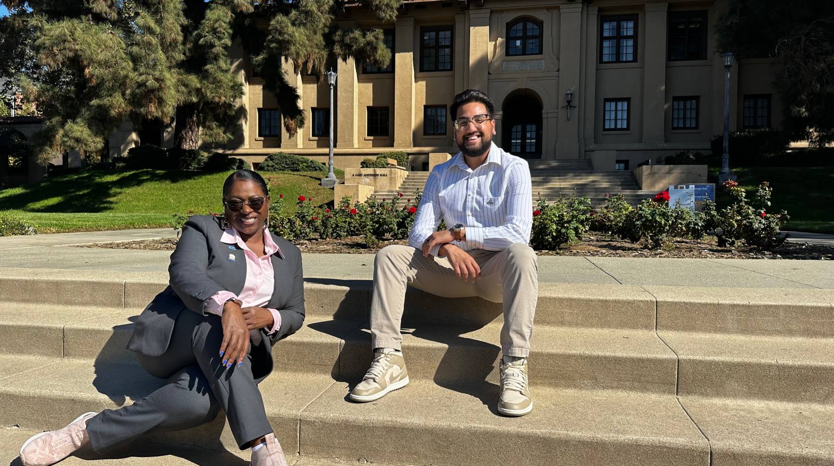 Sonya Brooks and Josiah Beharry, in business casual attire, sit on steps outside on a college campus and smile