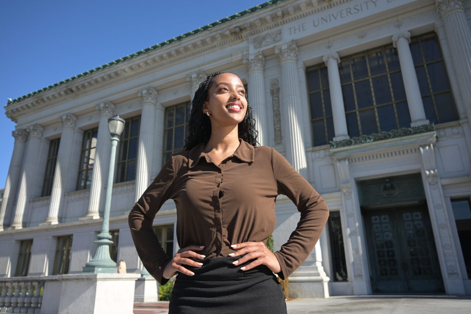 Woman with hands on hips looks off camera with a smile in front of a building on the Berkeley campus