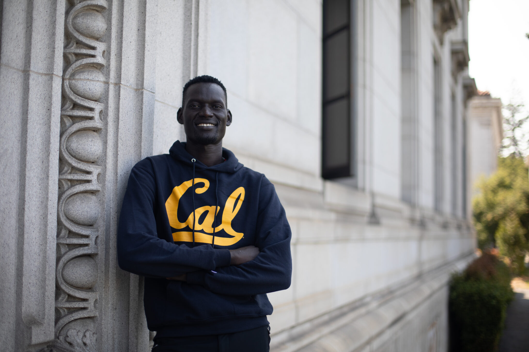 Young smiling man in Cal sweatshirt leans against a building on the UC Berkeley campus