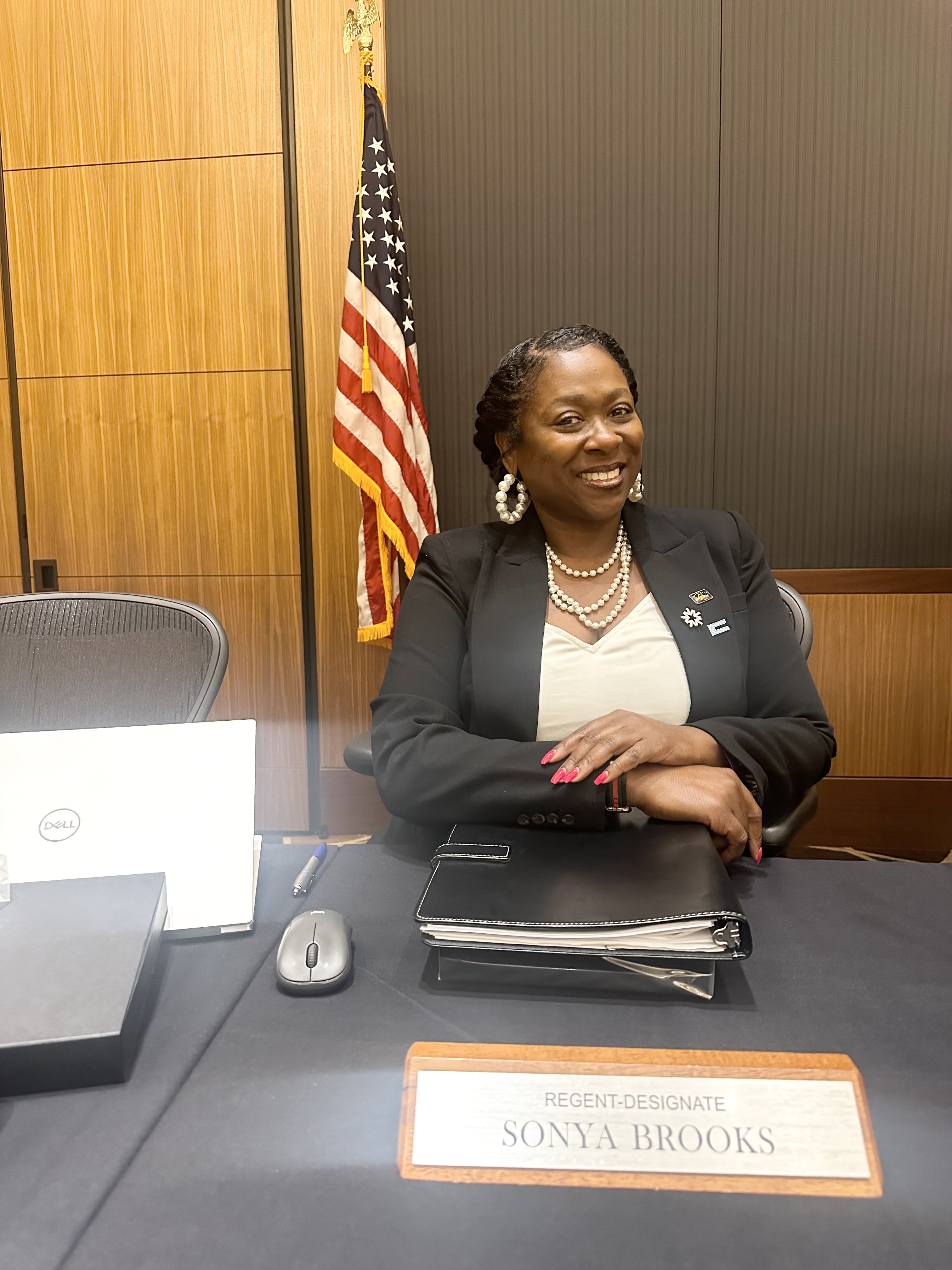 Sonya Brooks smiles at the camera at her spot around the UC Regent's meeting table, in front of her name placard.