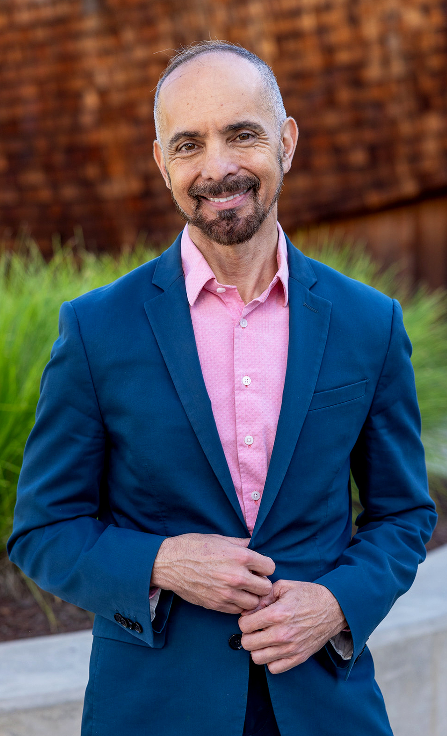 Senator Christopher Cabaldon smiles for a portrait, standing in front of a wood-shingled building with green plants in the background.