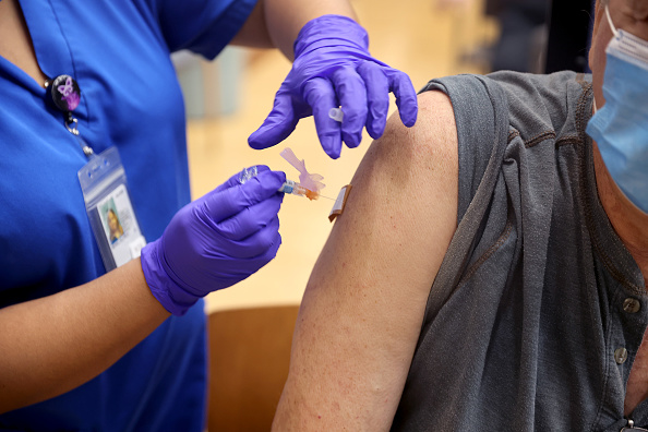 A woman in blue scrubs and purple gloves gives a shot to an older gentleman wearing a face mask.