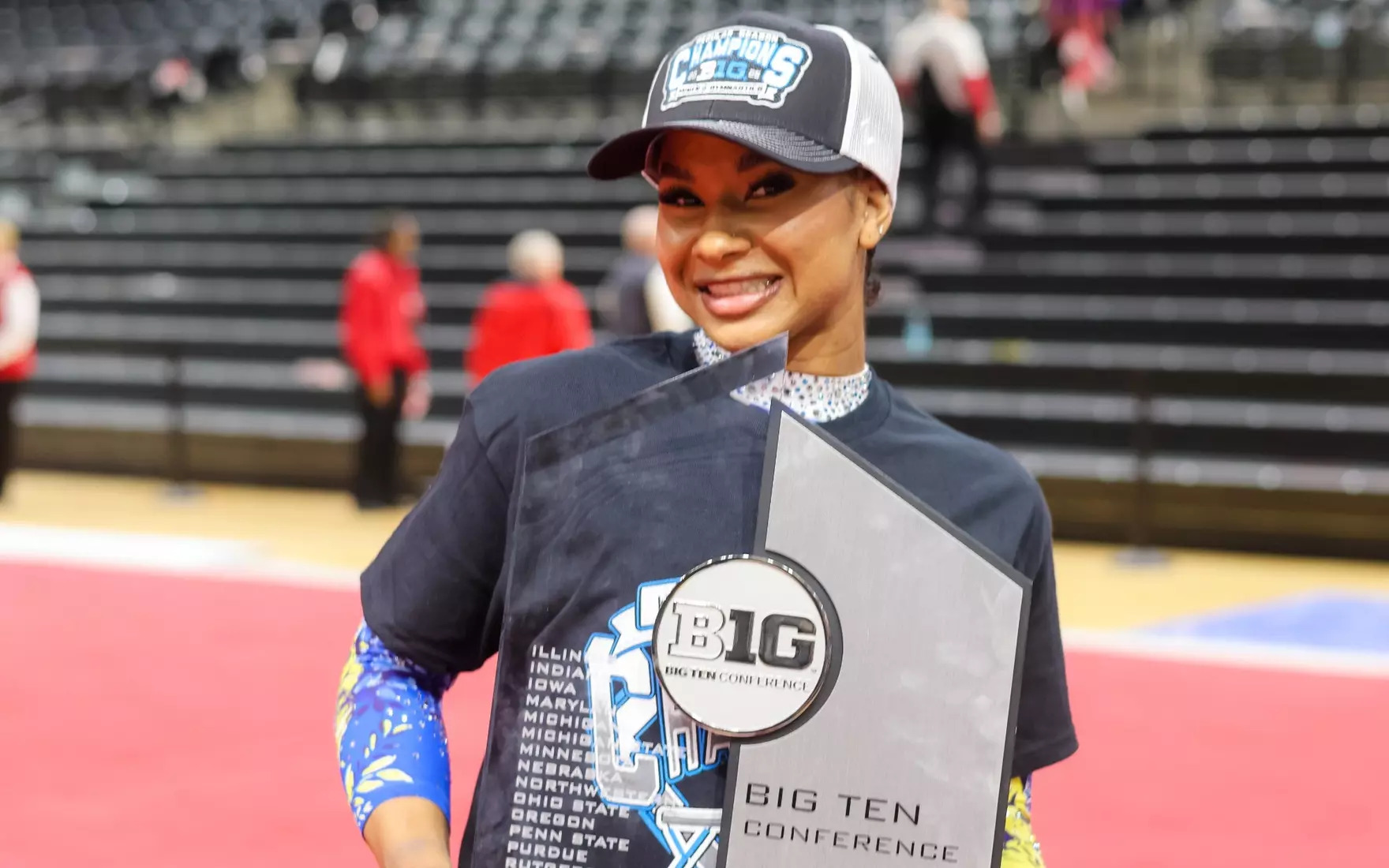 Jordan Chiles smiles, holding a Big 10 championship trophy and wearing a Big 10 champ baseball cap