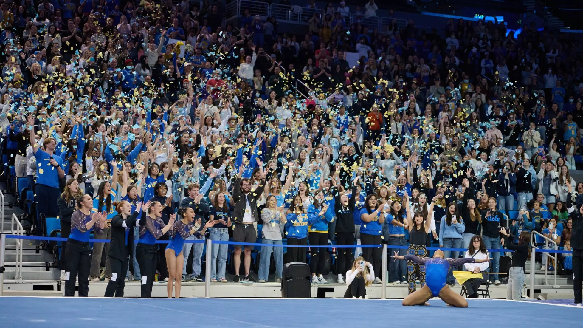 In the lower right corner, Jordan Chiles with her arms in the air on her knees after finishing a floor routine, confetti flying from the crowd as her teammates and the crowd screams at Pauley Pavilion