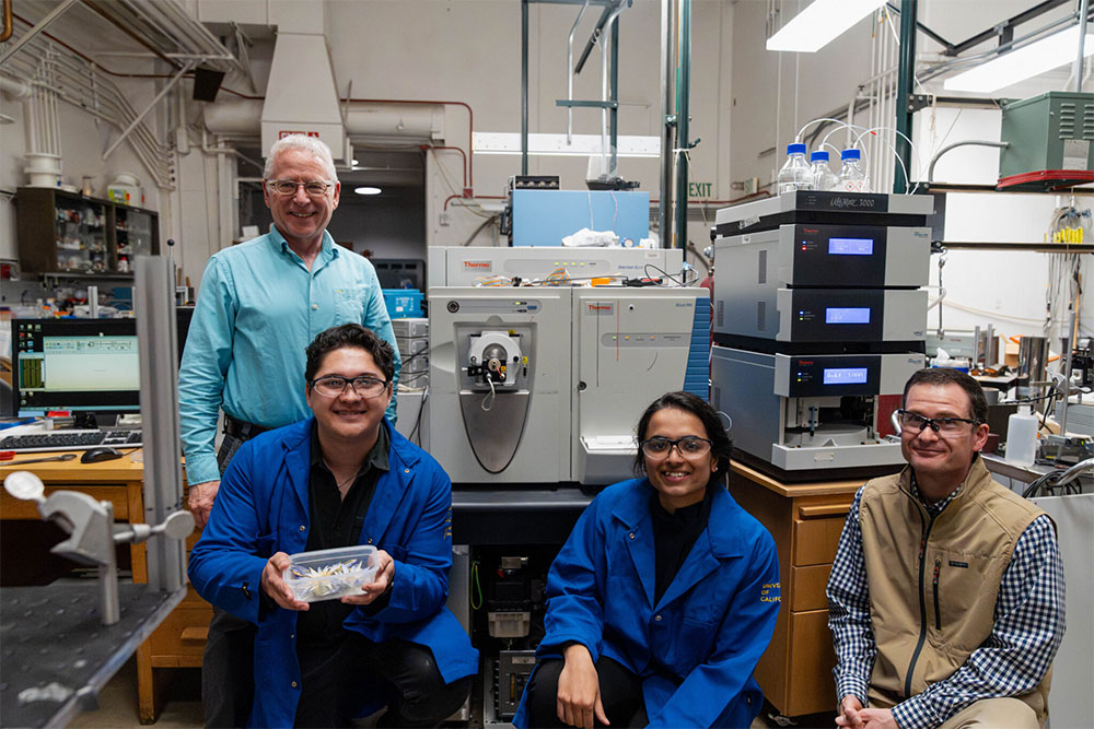 Four people in a lab, smiling for the camera, one holding a tray with several large flower blossoms in it