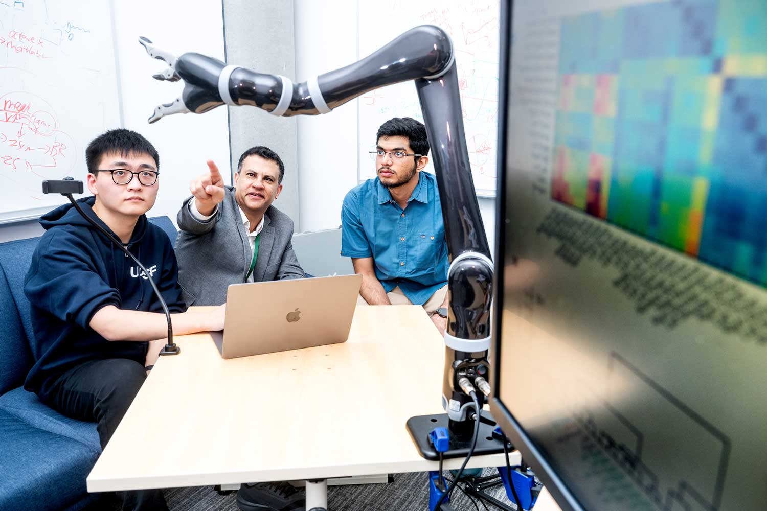 Three researchers sitting at a table with a laptop, looking at a robotic arm