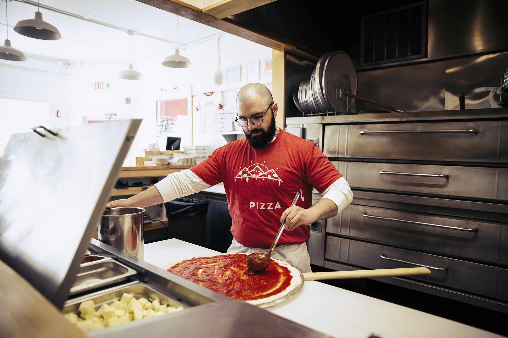 A chef puts pizza sauce on pizza dough in a commercial kitchen