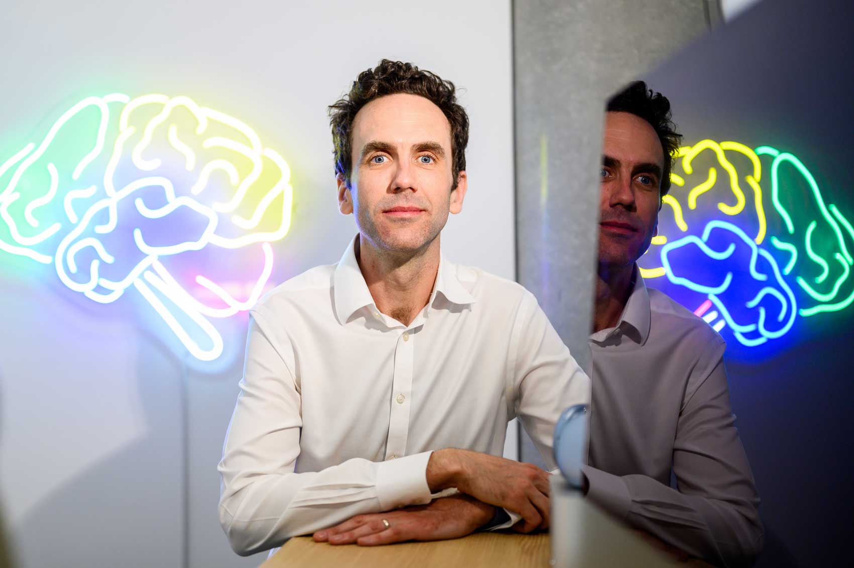 Simon Little poses for a portrait at a desk. Art on the wall behind him is a large shape of a brain rendered in neon lights.