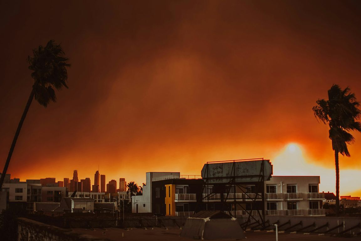 A dark orange sky with a palm tree, back of a billboard, and apartment complex in the foreground, Los Angeles skyline behind