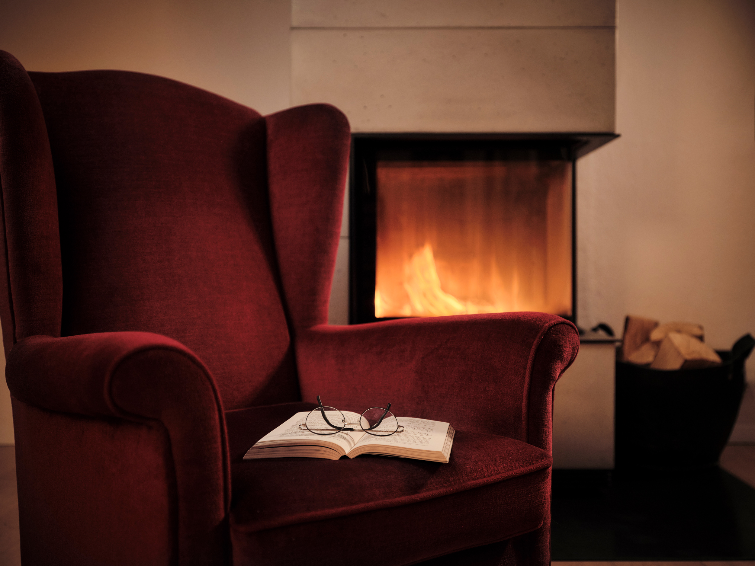 A deep red arm chair with an open book with glasses on top of it next to a fireplace