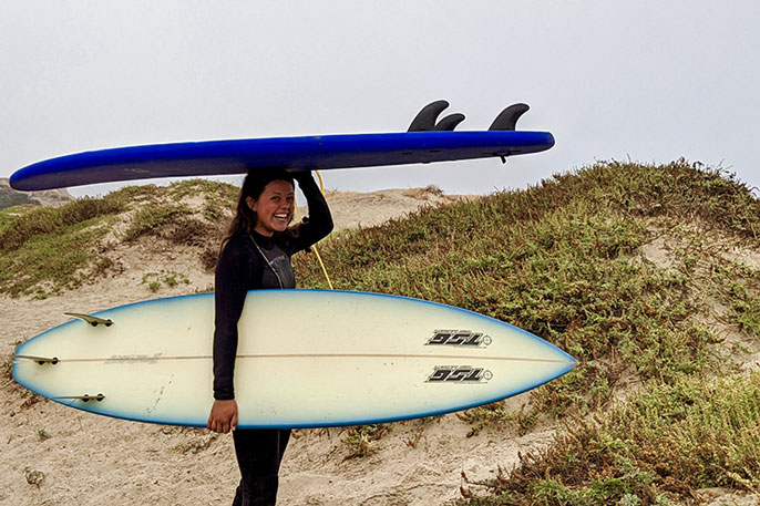 Katia Gibson, smiling, holding two surfboards on the beach on the way to the waves