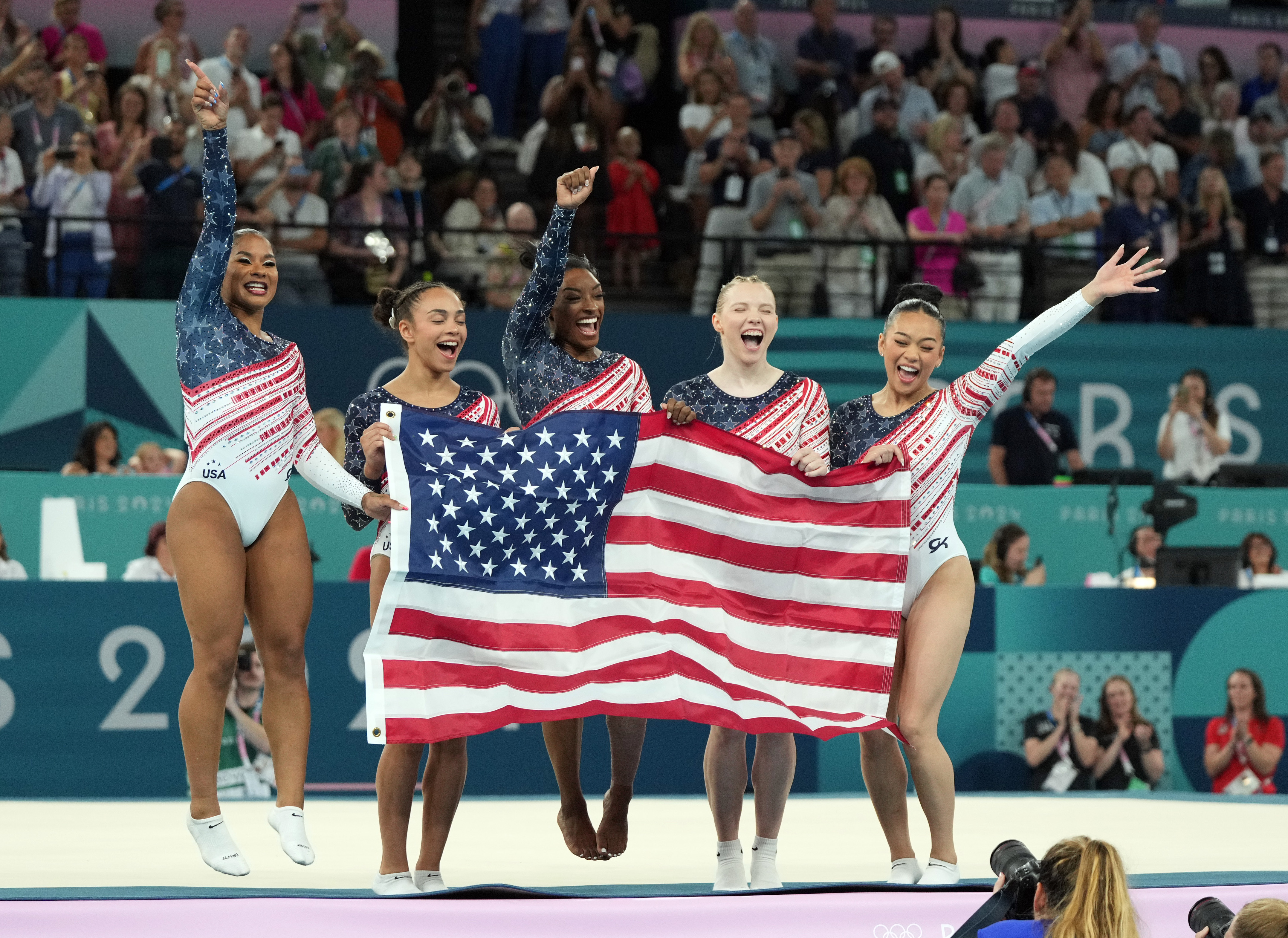 Five gymnasts from Team USA jump and scream while holding the American flag just after winning the 2024 Paris Olympics women's team competition