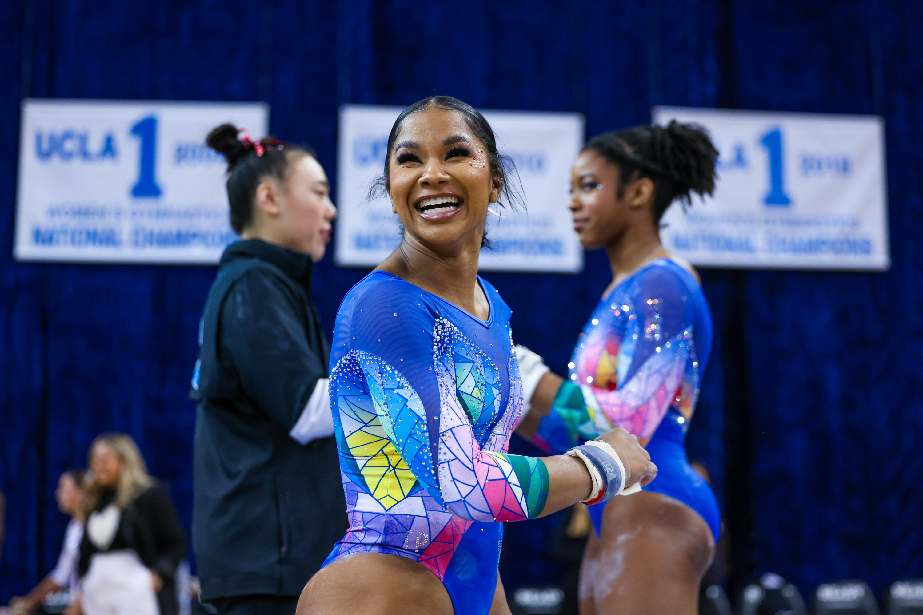 Jordan Chiles smiles while at a competition in a leotard, with another gymnast behind her on her right and a trainer behind her on her left