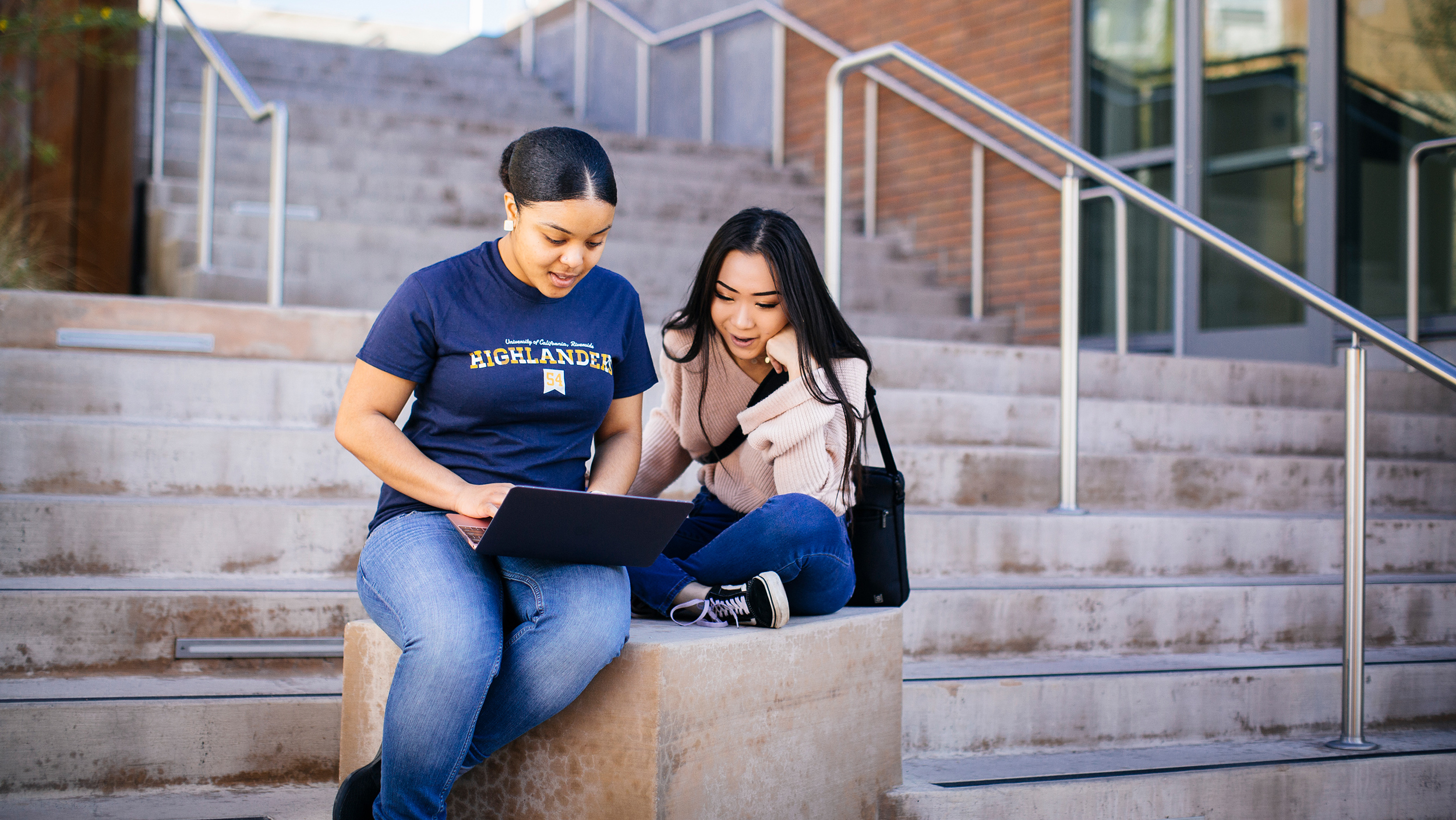 Two students sitting on a stairway landing outside looking at an open laptop