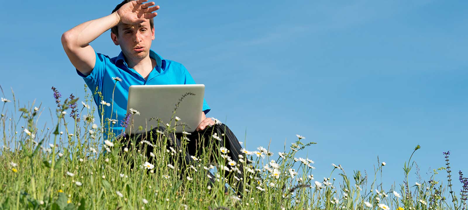 a man outside, sitting in a field with a laptop, wiping sweat off his brow