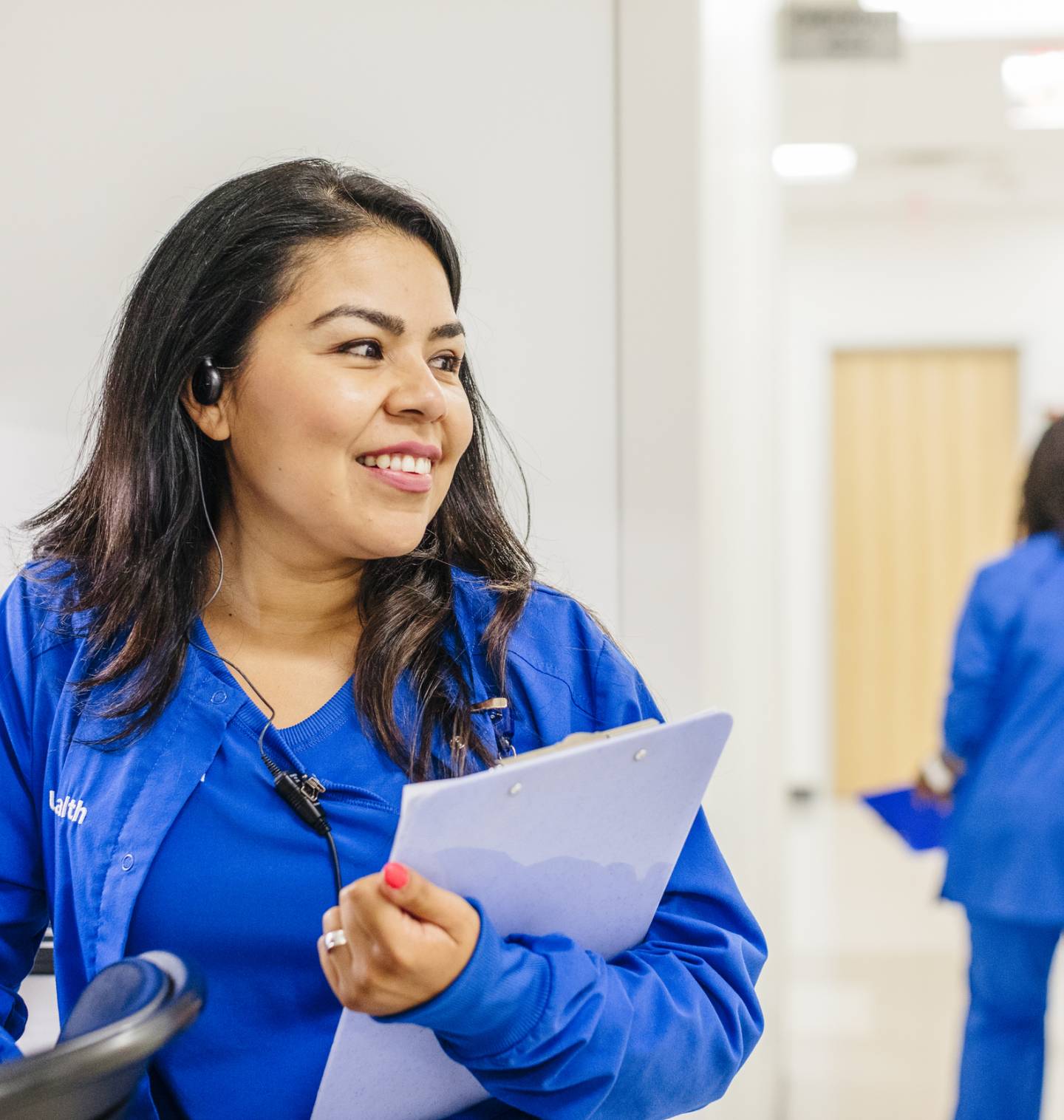 A smiling nurse in blue scrubs holds a chart as a patient and a nurse walk away behind her