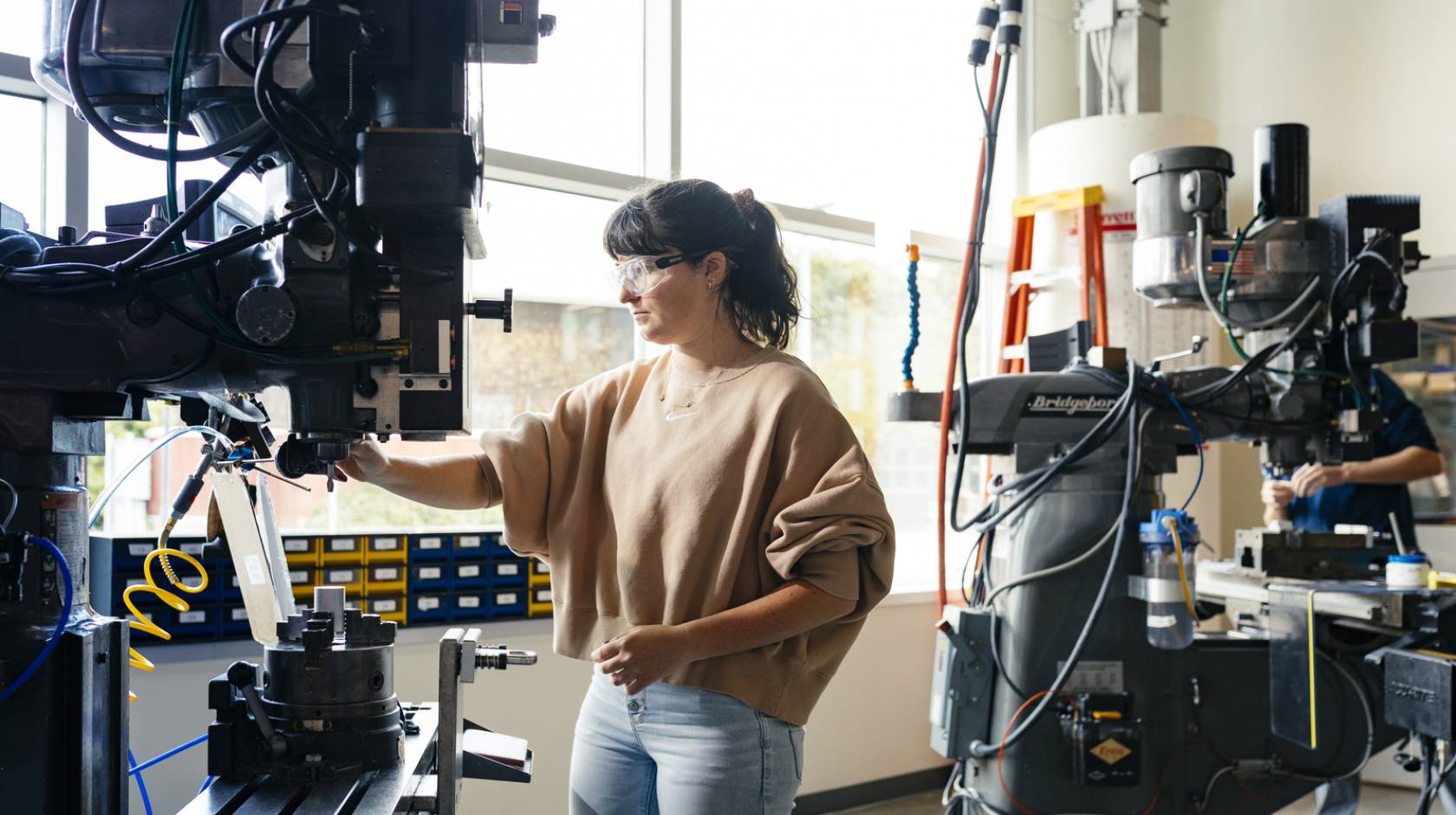 A woman in jeans and a brown sweater wearing safety glasses uses a large machine in a lab lit by natural light from a large wall of windows