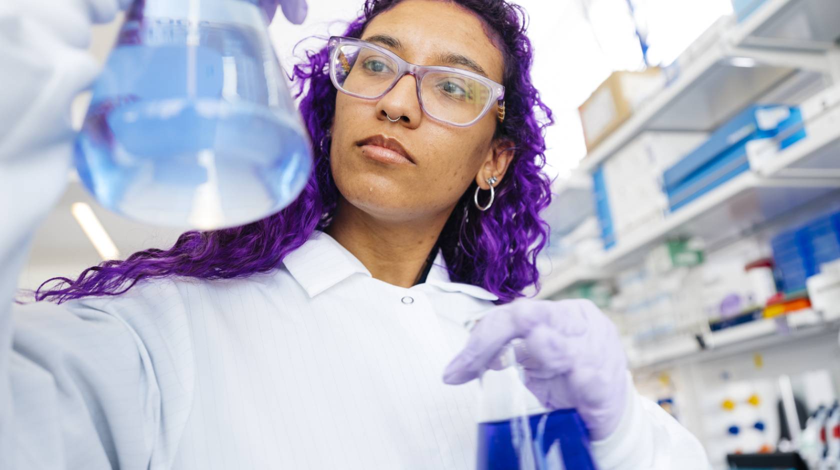 Woman with long, dyed-purple hair wearing a white lab coat and protective gear compares two flasks full of colorful liquid in a lab