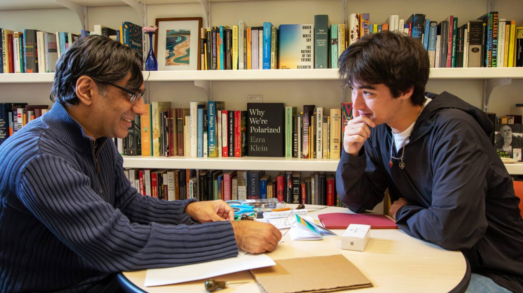 A professor-aged man and a student-aged man sit at a round table in front of a bookshelf, facing each other in discussion. 