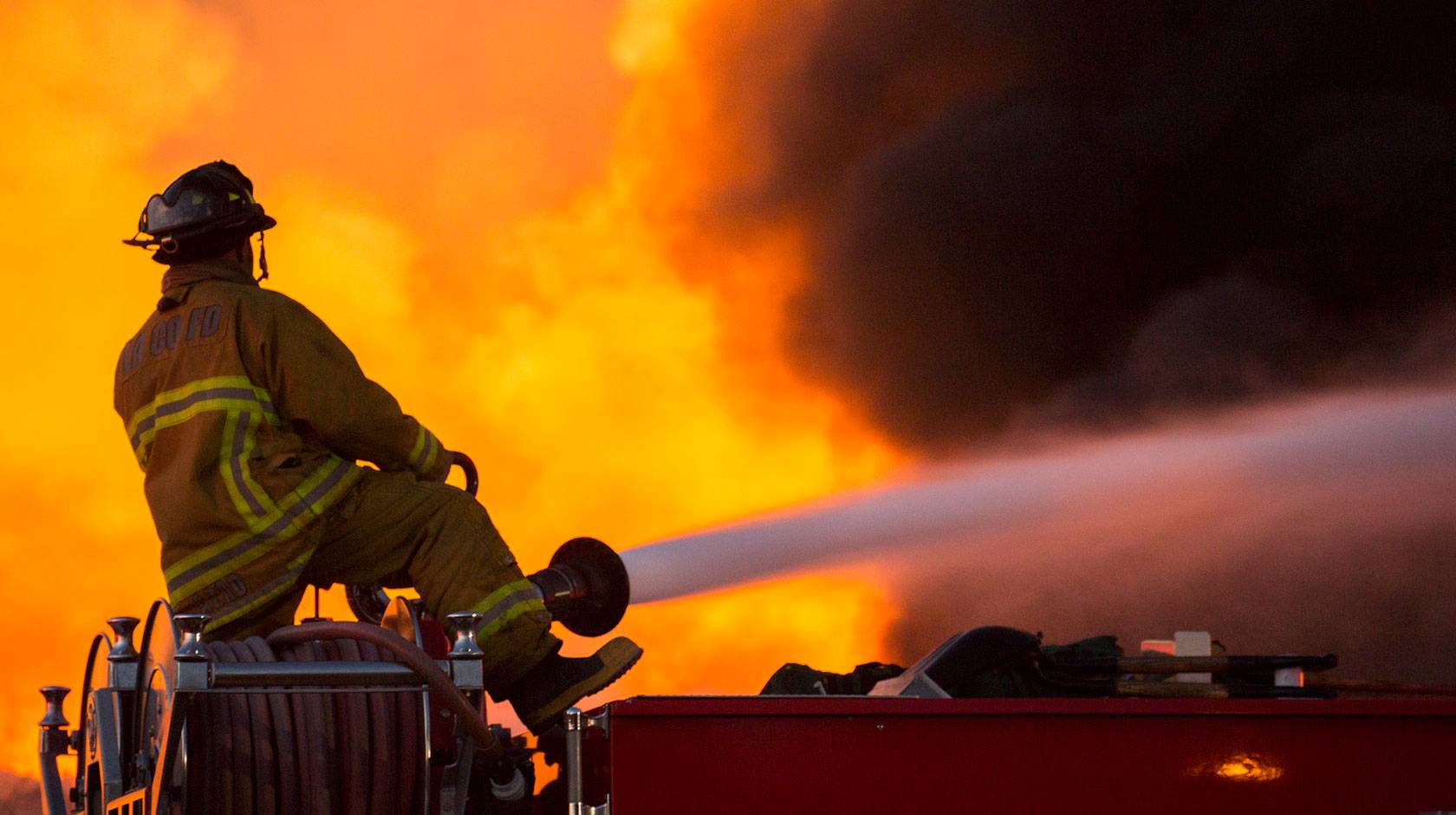 A firefighter straddles a roll of hose, aiming a jet of water into a wall of flames, facing away from the camera