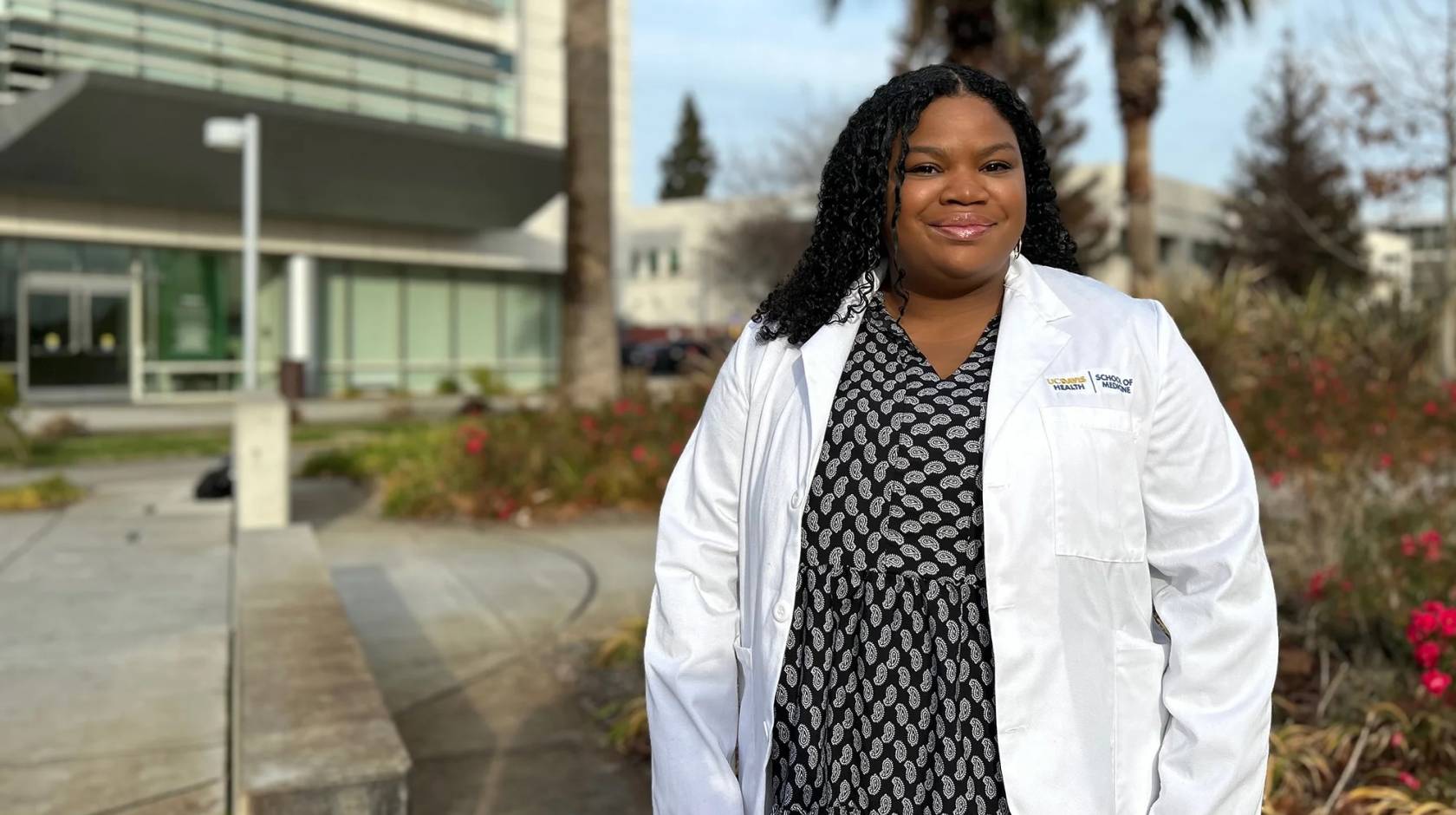 Chelsea Nash wears a black and white polka dot dress and white doctor coat, smiling for a portrait outside of a medical building at UC Davis