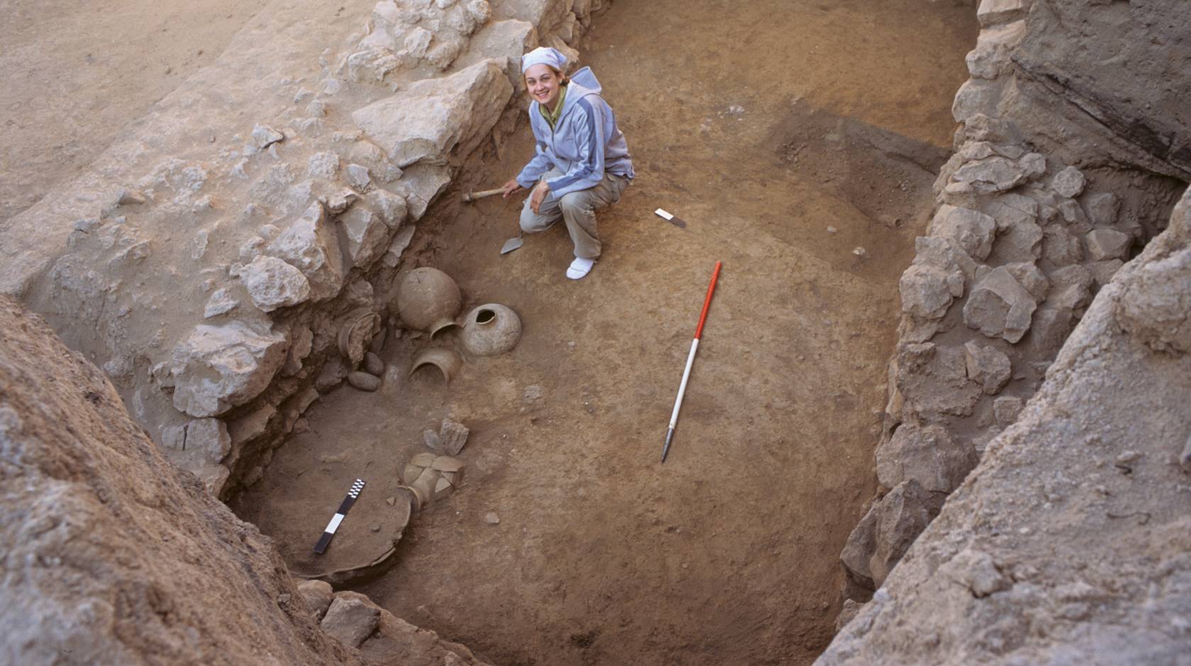 A woman in a very ancient, shallow tomb, looks up from her excavating work, smiling