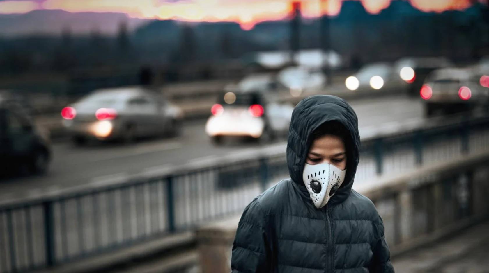 A boy in a hooded jacket standing next to a freeway wearing a respirator mask