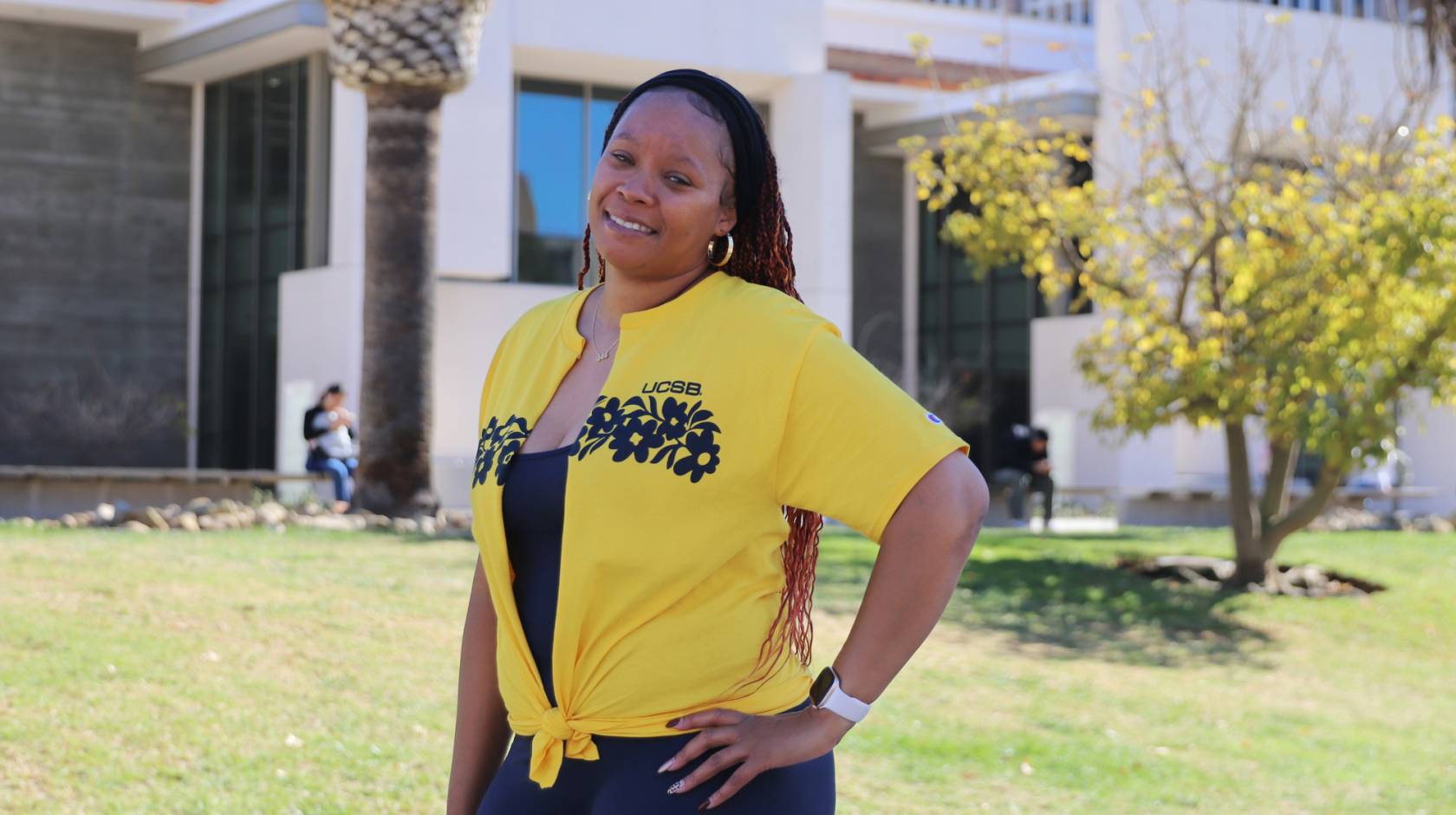 A smiling woman in a yellow UCSB shirt standing in front of a building on campus