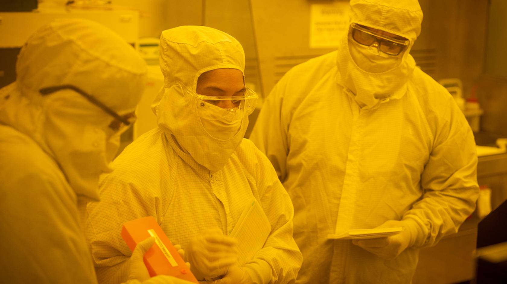 Three people stand in white full-body suits in the yellow light of Nano3's cleanroom.