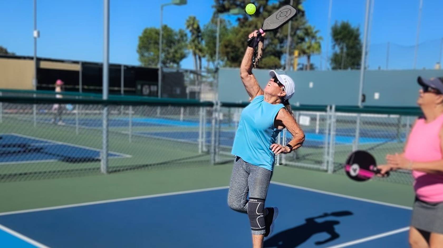 Two women on a pickleball court. A woman in the center of the frame reaches to hit a ball overhead, and her teammate looks on