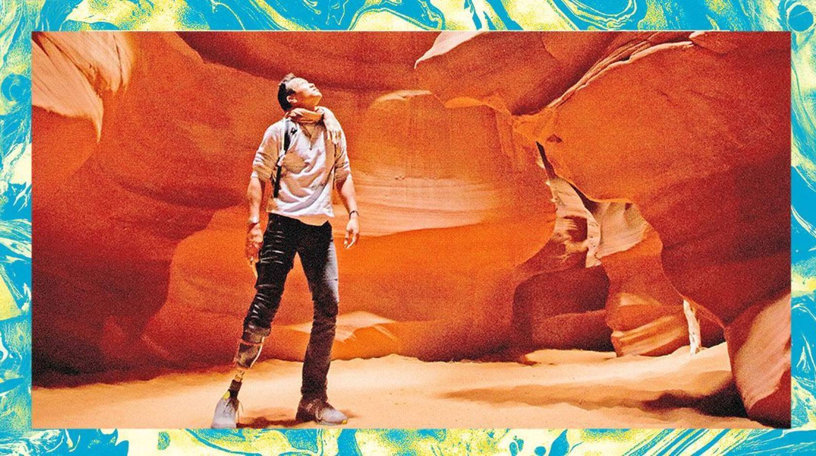 A male hiker with a prosthetic leg below his knee looks up against the backdrop of a red rock canyon wall. The photo is edited and filtered to look grainy, and surrounded by a teal/green textured frame.