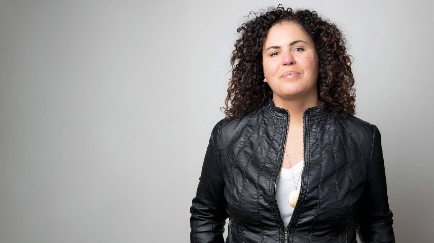 Safiya Noble, wearing a cool black leather jacket, looks resolute in a waist-up portrait against a gray background