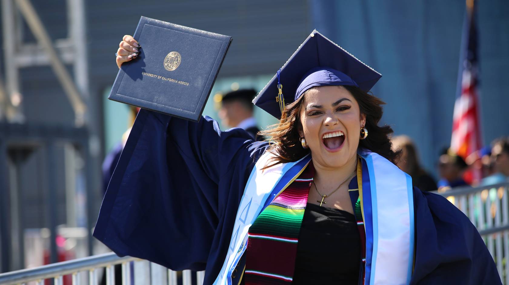 A woman in a cap and gown smiles while holding up her diploma